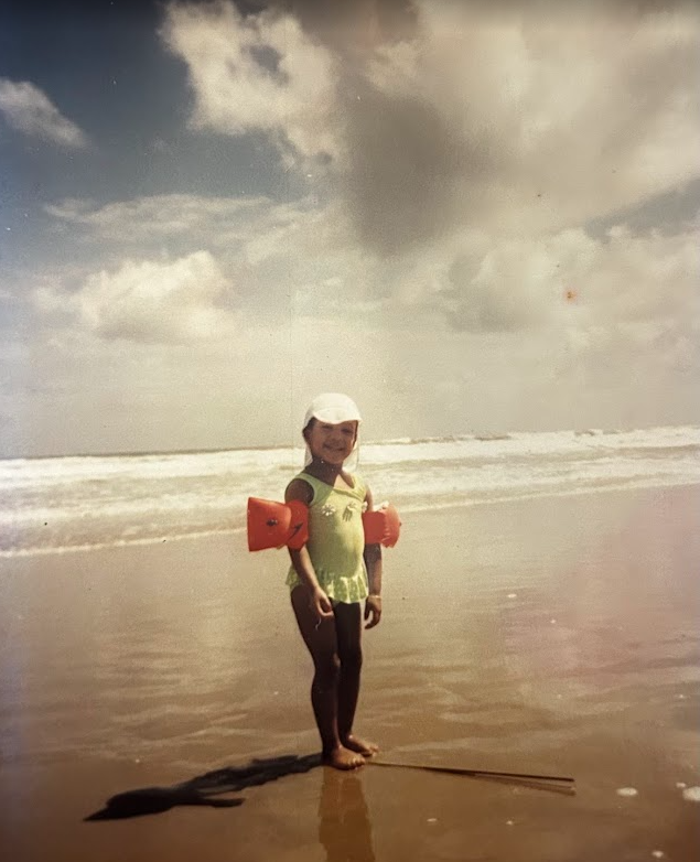 Young Tsian standing at the water’s edge on a beach, wearing a green swimsuit and orange arm floats.